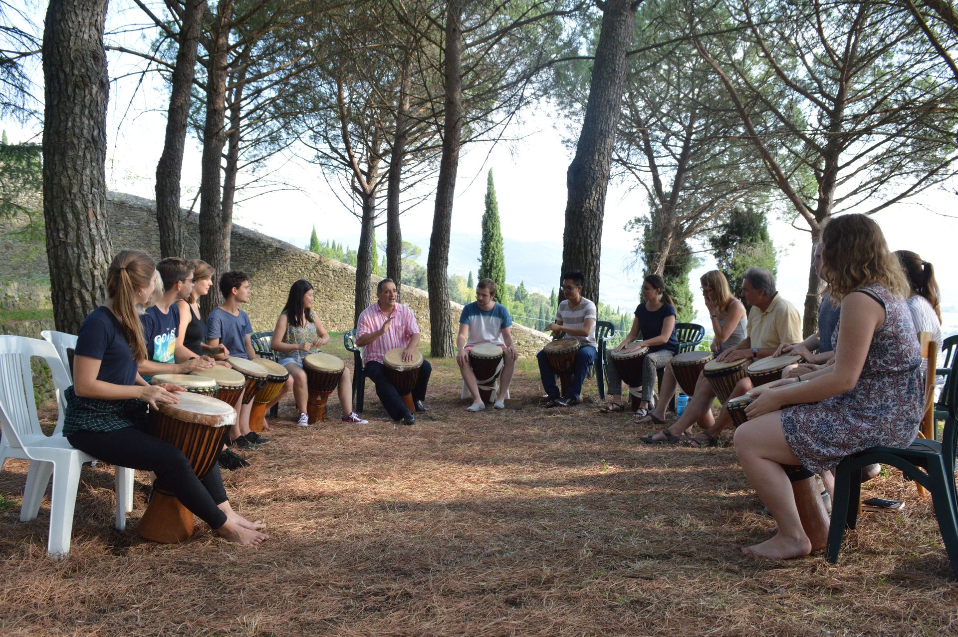 Participants play hand drums together in a circle beneath the pine trees.
